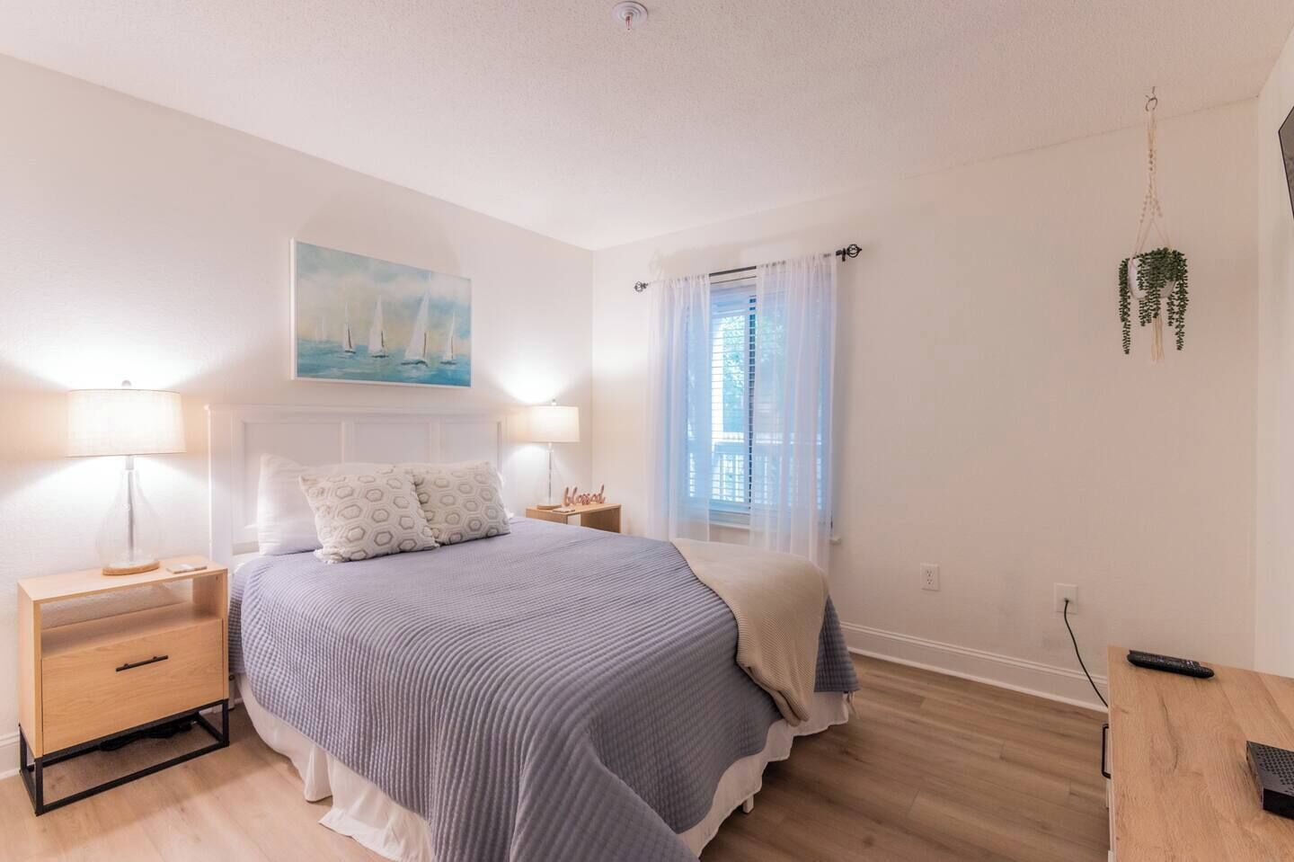Un dormitorio con cama, mesitas de noche, un cuadro, una ventana con cortinas y una planta colgante.