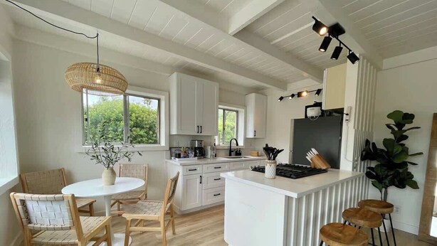 A modern kitchen with white cabinetry, a dining area with wooden chairs, and a bar with stools. - Encinitas Surf Shack w/ Private Rooftop Oasis (Encinitas)