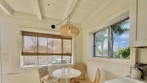 A room with a round table, two woven chairs, a hanging light fixture, a window with blinds, and a view of palm trees. - Encinitas Surf Shack w/ Private Rooftop Oasis (Encinitas)