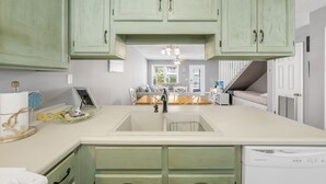 A kitchen with green cabinets, a white countertop, and a dishwasher. There is a dining area with a table and chairs in the background. - Emerald Coast Paradise Condo, Steps from PCB Beaches! (Panama City Beach)