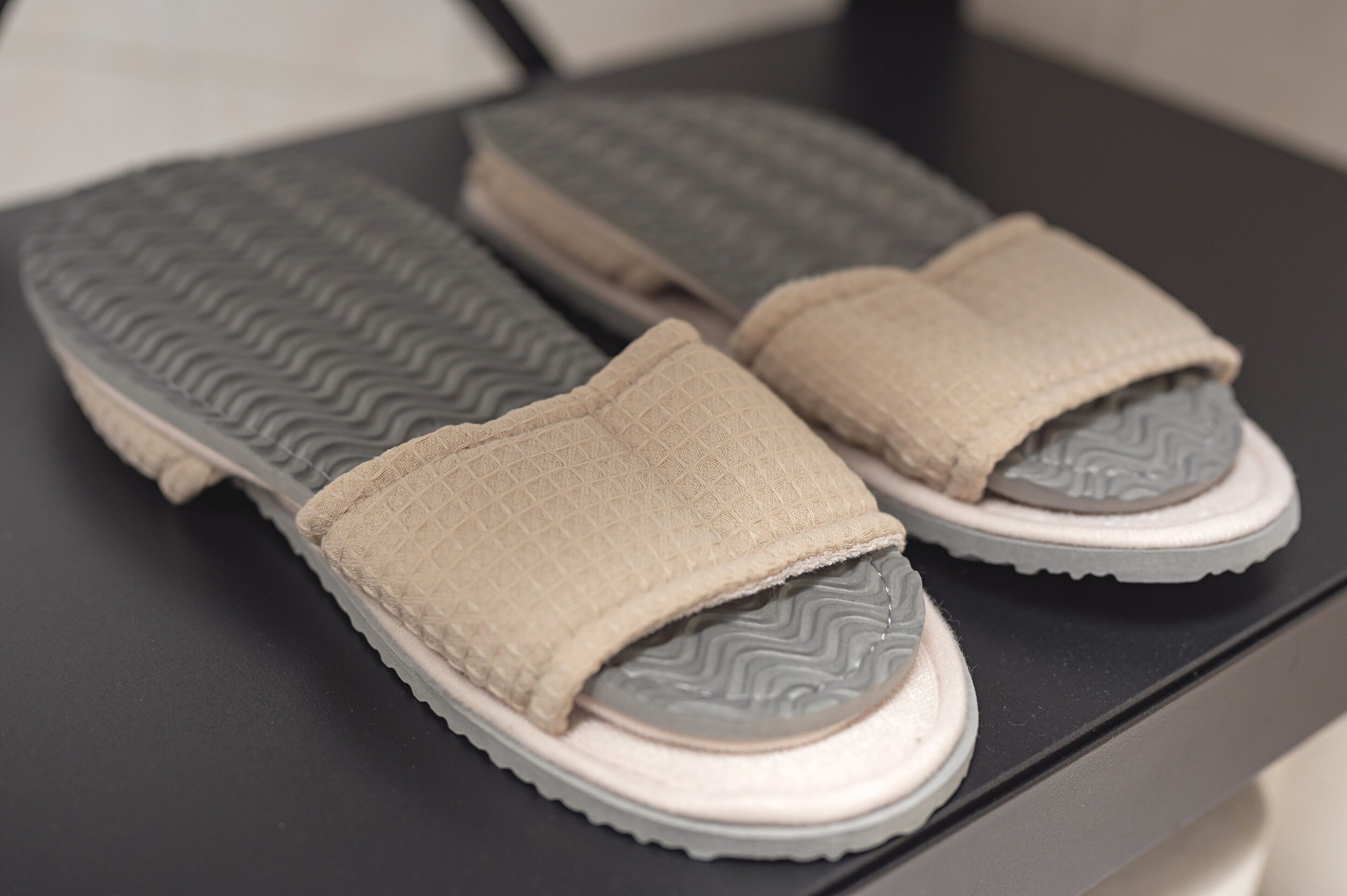 Une paire de pantoufles beiges à semelle texturée, posées sur une surface noire.