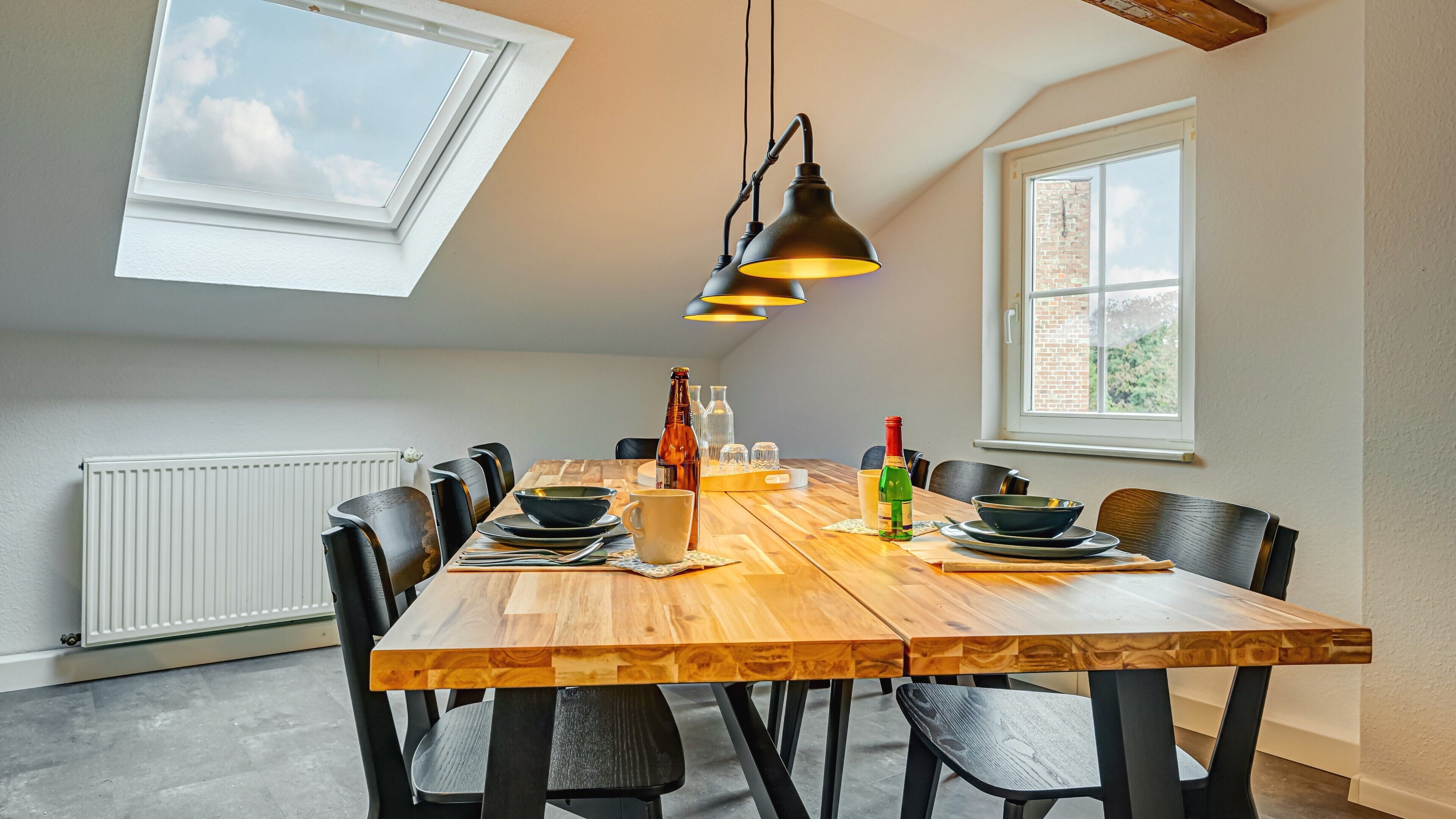 Un coin repas avec une table en bois, des chaises noires, une bouteille, des verres et une fenêtre.