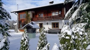 Un chalet a due piani con un balcone in legno, un piccolo deposito annesso e un paesaggio innevato.