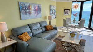 Un salon moderne avec un canapé en cuir gris, une table basse et un coin repas comprenant des chaises et une table. Les murs sont ornés de tableaux abstraits et de grandes fenêtres offrent une vue sur l’océan.