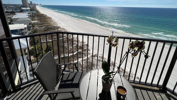 Un balcon avec vue sur une plage et l’océan. On y trouve une chaise, une petite table avec une plante et une tasse, ainsi qu’une rambarde.