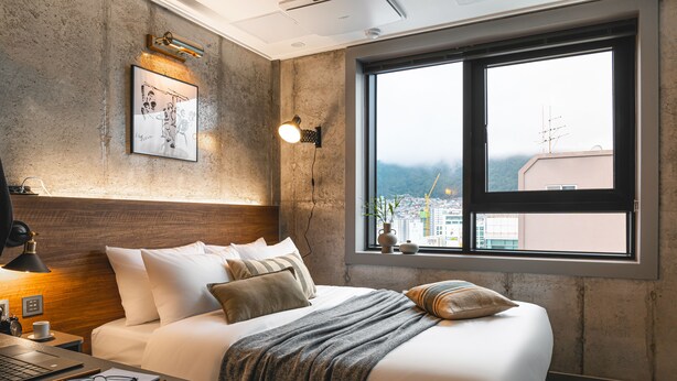 Una camera d'albergo moderna con un letto grande, una scrivania con un portatile, una lampada a parete e una finestra con vista sulla città.