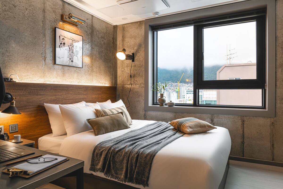 Une chambre d’hôtel moderne avec un grand lit, un bureau avec un ordinateur portable, une lampe fixée au mur et une fenêtre donnant sur la ville.