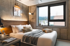 A modern hotel room with a large bed, a desk with a laptop, a wall-mounted lamp, and a window with a city view. - Urbanstay Boutique Nampo BIFF (Busan)