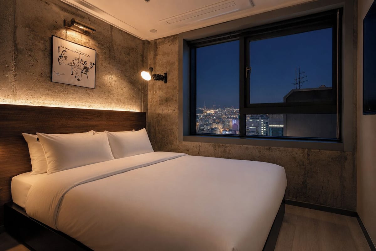 Une chambre d’hôtel avec un grand lit, une vue sur la ville par la fenêtre et un luminaire fixé au mur.