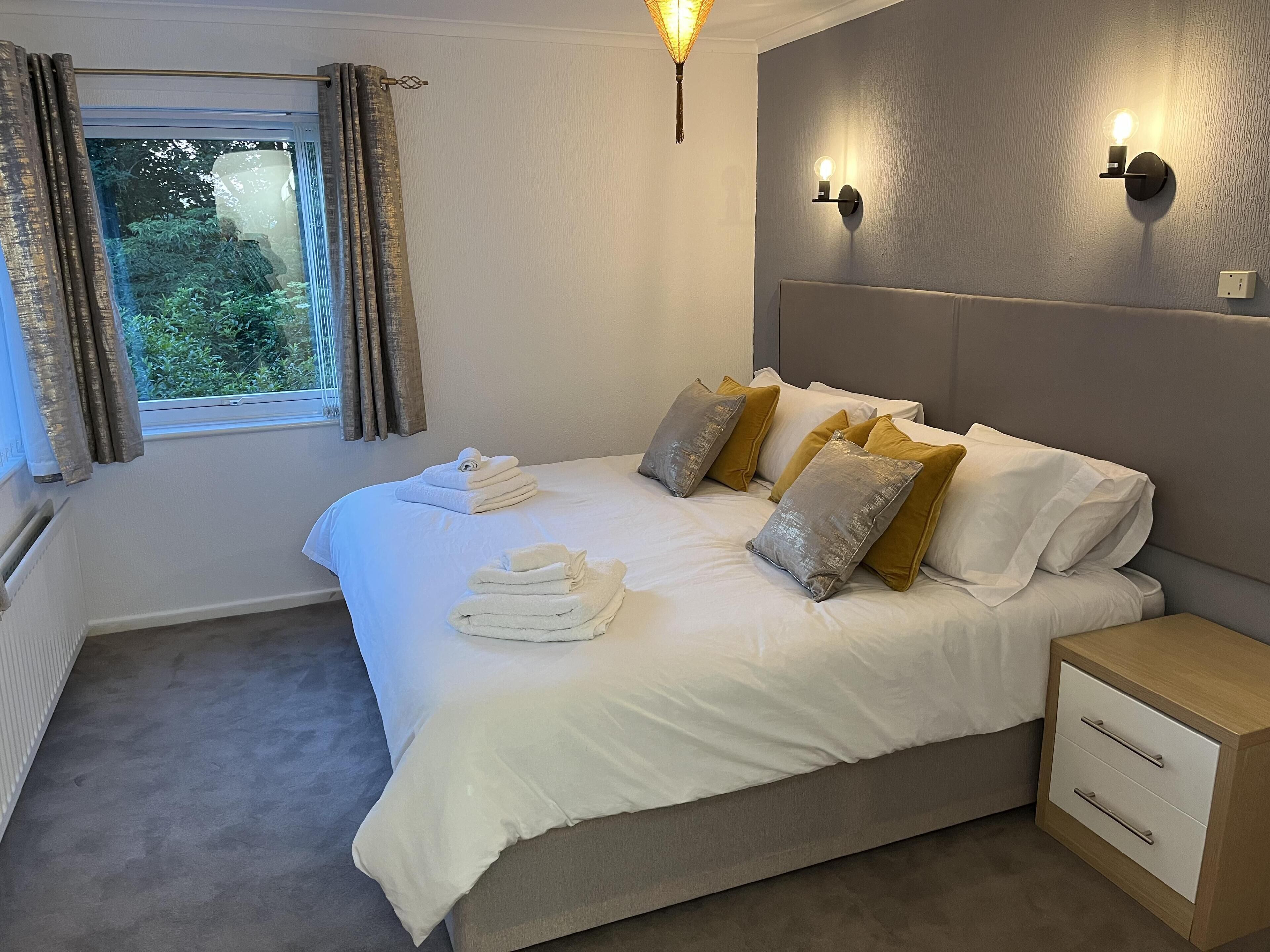 A neatly made bed with white and gold pillows, a bedside table with drawers, and a window with curtains.