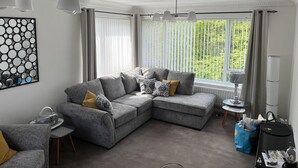 A modern living room with a grey sectional sofa, a small round table, and a large window with vertical blinds. - Tranquil Retreat on the Edge of the Countryside (Blaydon-on-Tyne)