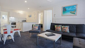 A modern living room with a sofa, coffee table, and chairs. There is a kitchen area with a microwave and a dining table. A painting of a boat is on the wall.