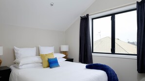 Habitación de hotel con una cama grande, ropa de cama blanca, una manta azul y vistas de edificios a través de una ventana con cortinas negras.