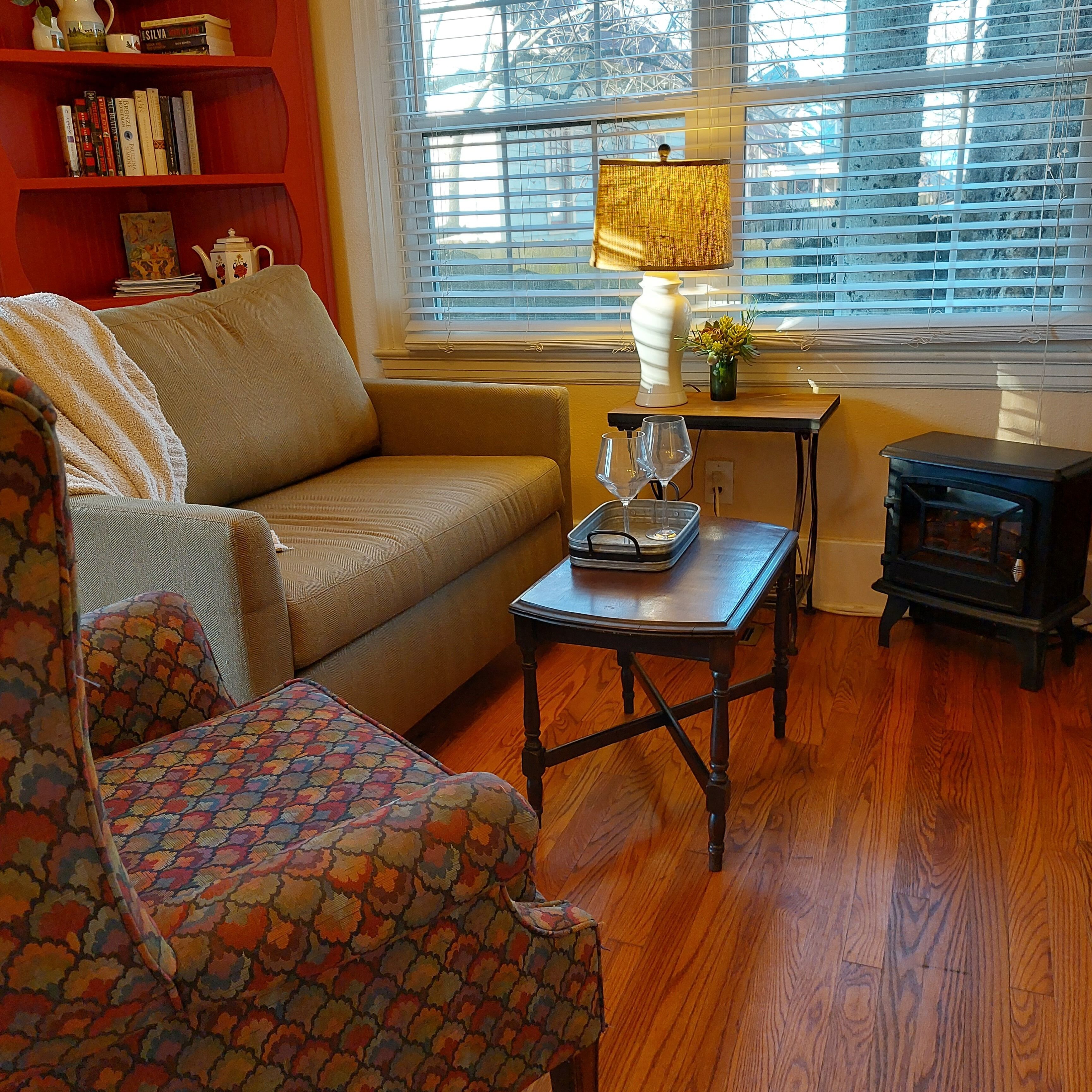 A cozy living room with a sofa, a patterned armchair, a small table with a tray and glasses, a lamp, and a bookshelf.