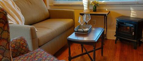 A cozy living room with a sofa, a patterned armchair, a small table with a tray and glasses, a lamp, and a bookshelf.
