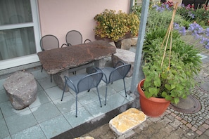 Un espace extérieur aménagé avec une table en pierre et des chaises en métal, entouré de plantes en pot et doté d’un sol carrelé.