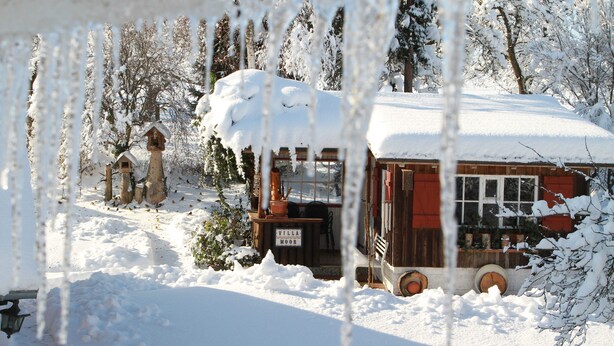 A snow-covered wooden cabin with a sign reading 'Villa Moor,' surrounded by trees and a snow-covered yard.