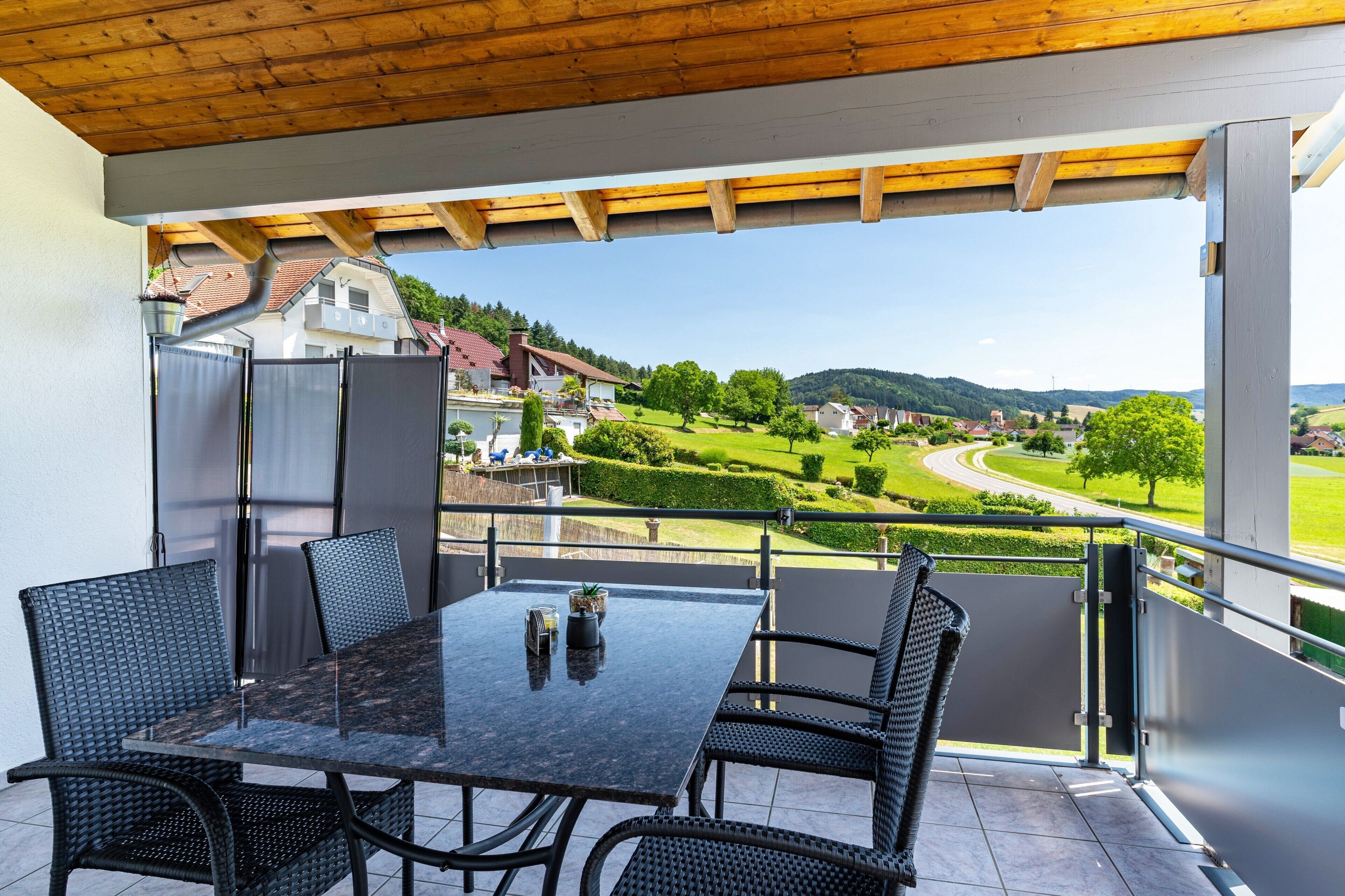 Een balkon met een tafel en stoelen, een uitzicht op een groen landschap, en een gebouw op de achtergrond.
