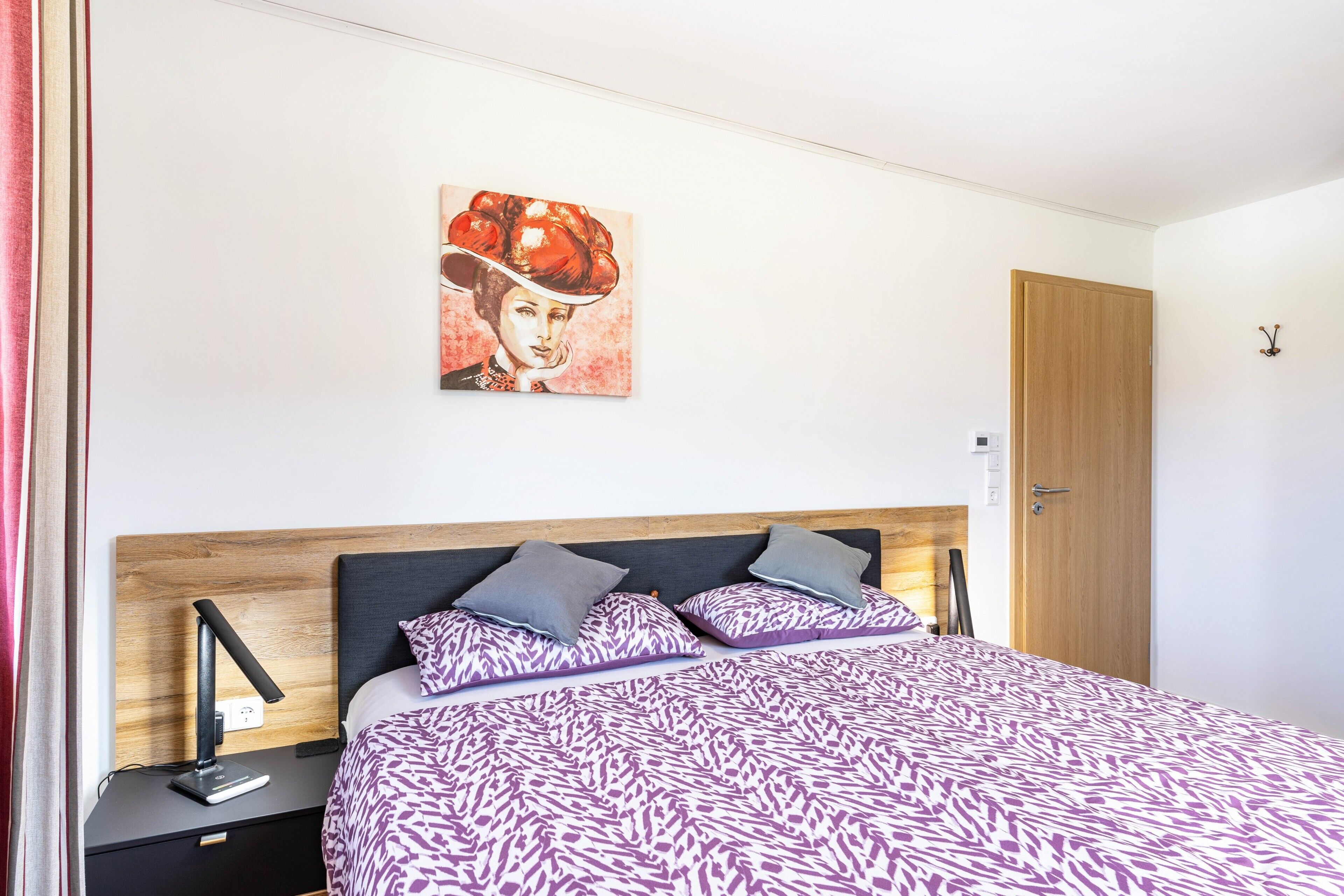 A bedroom with a patterned bedspread, a nightstand with a lamp, a wall-mounted painting, and a wooden door.