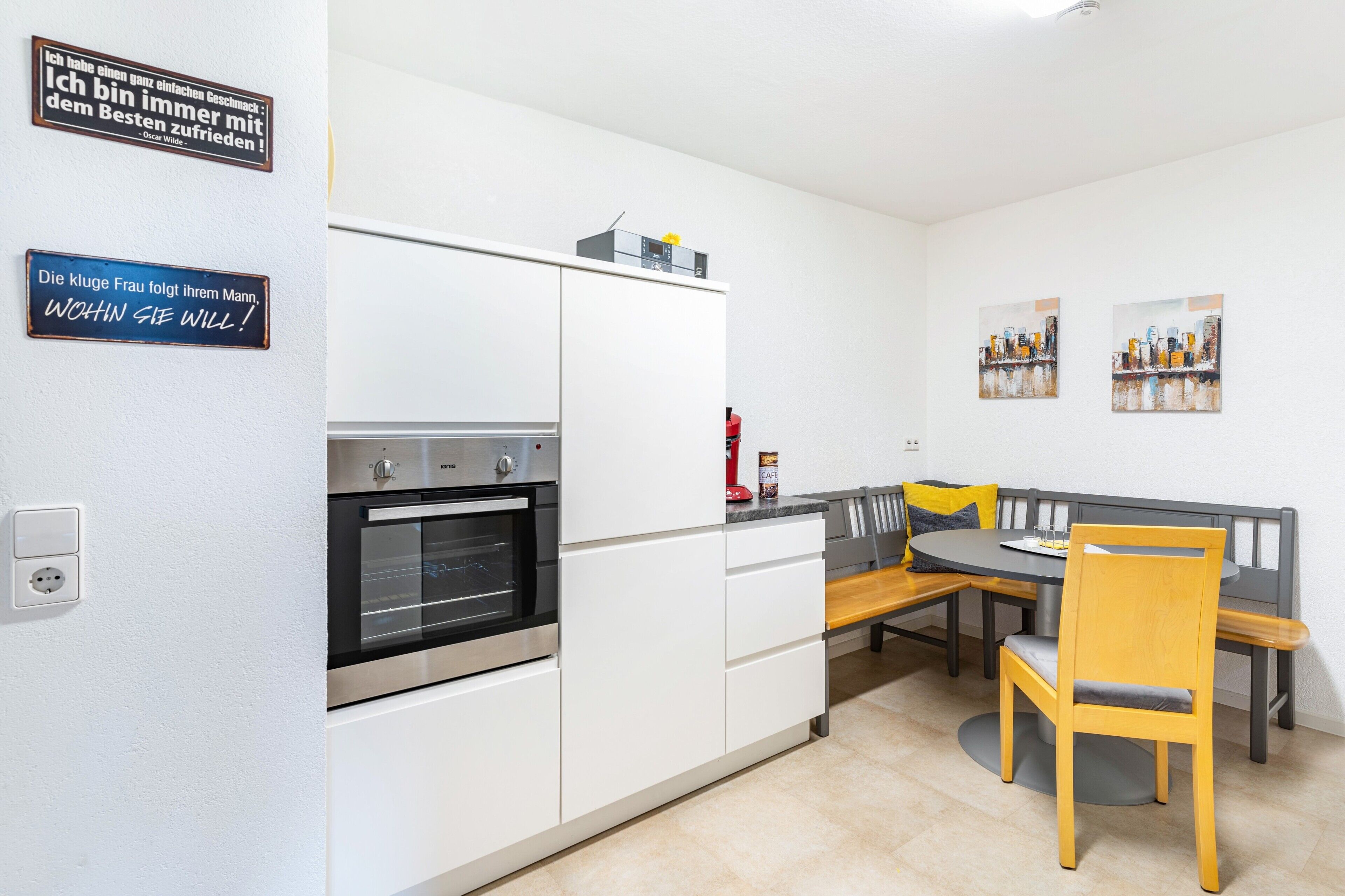 Une cuisine moderne avec des meubles blancs, un four encastré et un coin repas comprenant une banquette et une chaise.