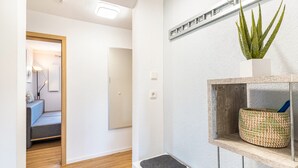 Ein Flur mit Holzboden, einem grauen Sofa, einer weißen Wand, einem Regal mit Pflanze und Korb sowie einer Tür zu einem weiteren Zimmer.