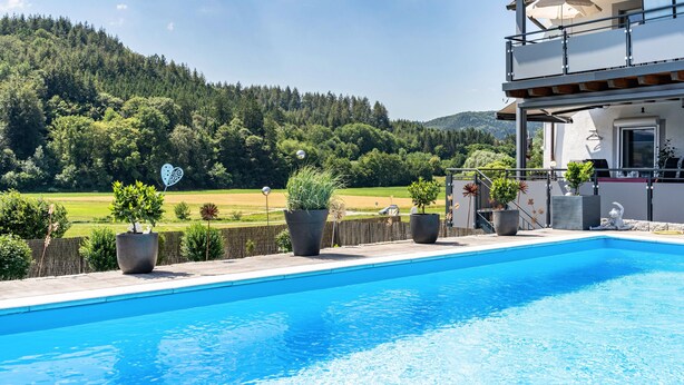 Ein Swimmingpool mit Blick auf einen bewaldeten Hügel, ein Balkon mit einer Pergola und Kübelpflanzen.