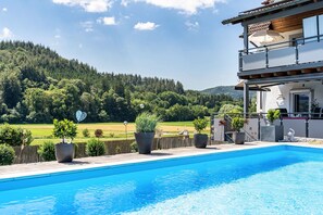 Una piscina con vistas a una ladera boscosa, un balcón con pérgola y plantas en macetas.