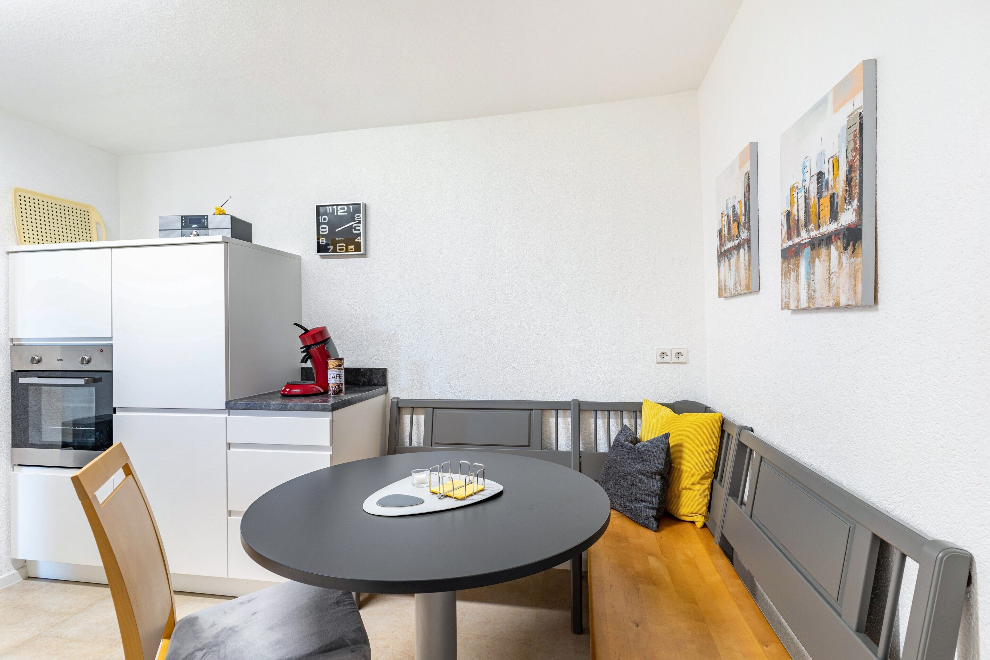 A modern kitchen with a dining area, including a round table with a tray, a bench with cushions, and a refrigerator.