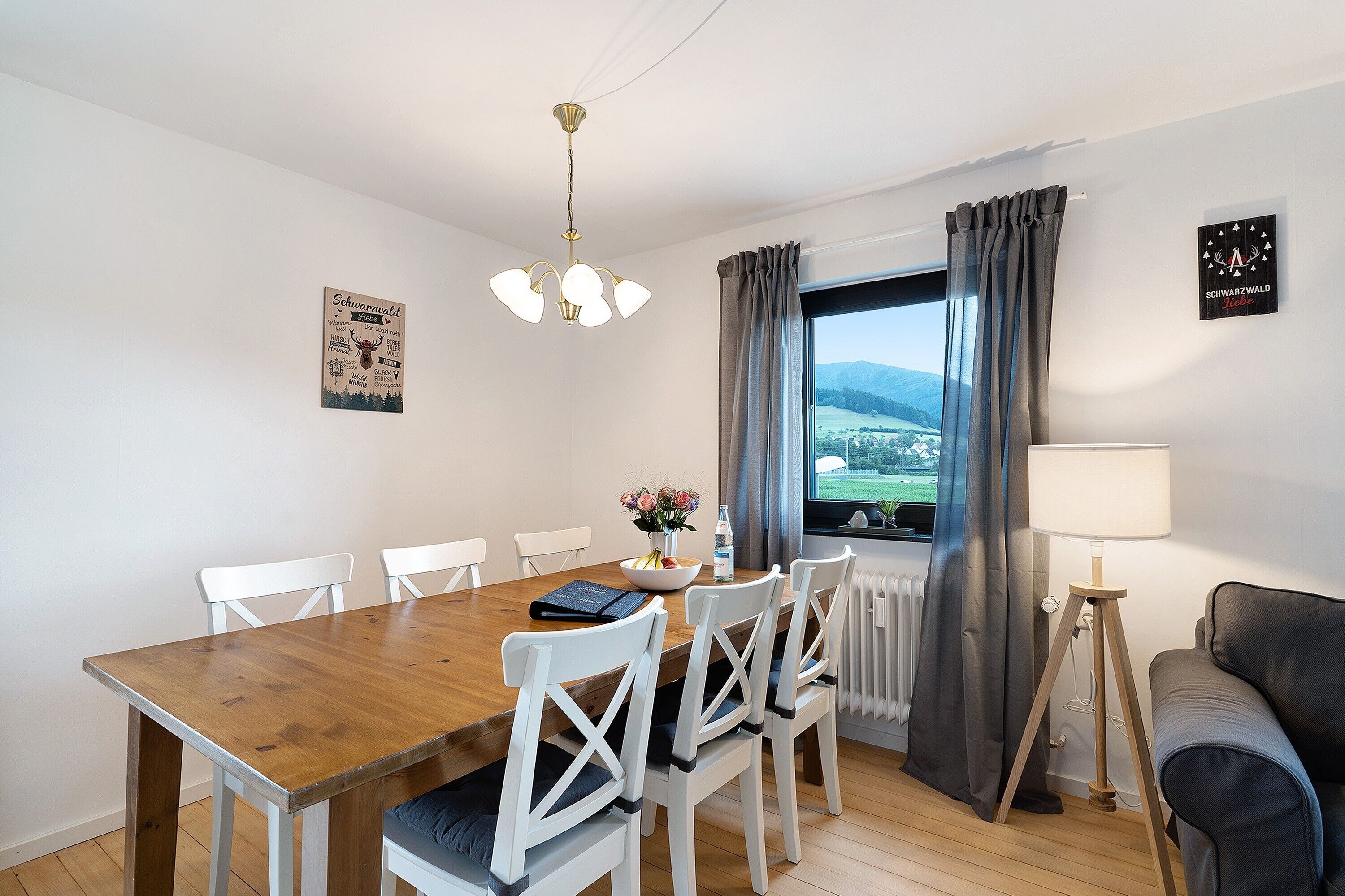 Ein Essbereich mit Holztisch, weißen Stühlen, einer Chandelüster, Blick durch ein Fenster und einer Lampe.