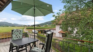Un balcón con mesa y sillas, un paraguas verde y vistas a un paisaje rural con casas y campos.