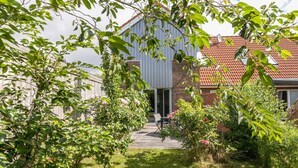 Ett hus med en fasad av tegel, en träveranda och en trädgård med olika växter och blommor.