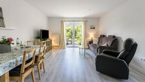 Ett kompakt vardagsrum med ett matbord, stolar, en soffa, en fåtölj, en TV och en skjutdörr i glas som leder ut till ett utomhusområde.