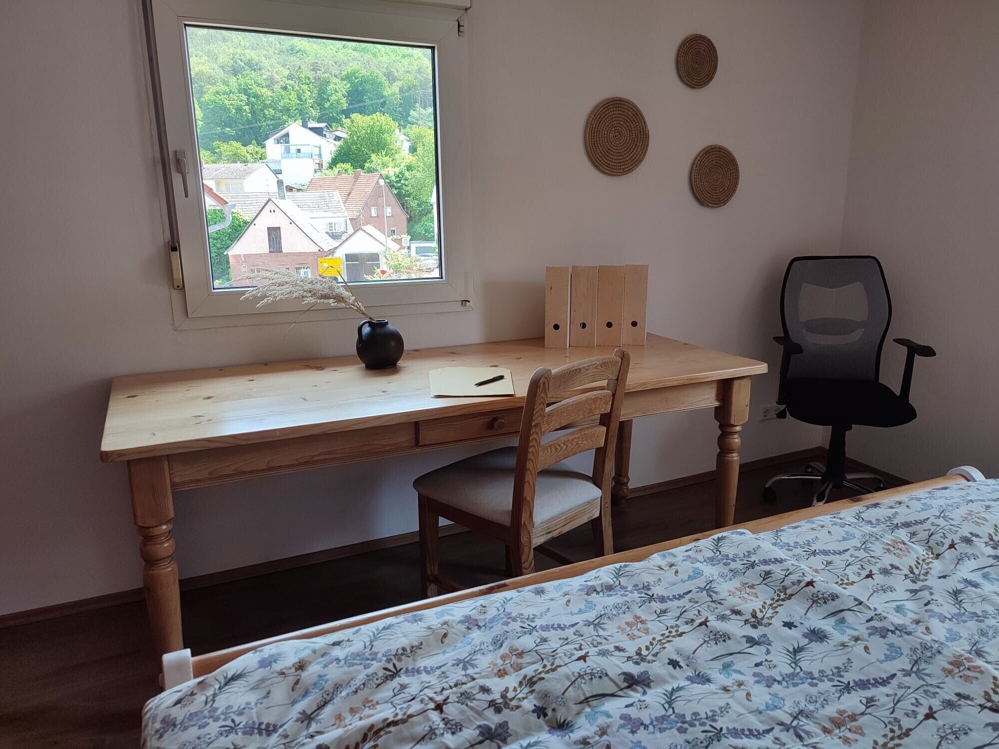 A room with a wooden desk, a chair, a bed with a floral bedspread, a window with a view of houses, and three wall decorations.