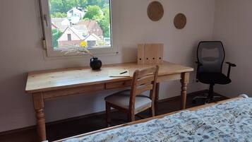 A room with a wooden desk, a chair, a bed with a floral bedspread, a window with a view of houses, and three wall decorations.