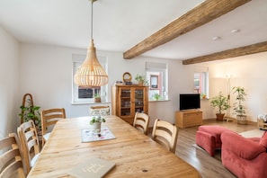 A dining area with a wooden table, chairs, a hanging light fixture, a cabinet with glassware, and a television.