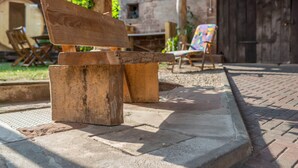 Un banc en bois avec un dossier, une table en bois et une chaise colorée, le tout installé dans un espace extérieur avec un mur de briques et de la verdure en arrière-plan.