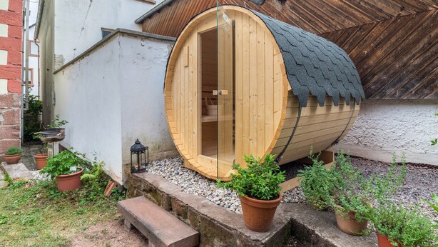 Une structure de sauna en bois dotée d’une porte en verre, entourée de plantes en pot et agrémentée d’un banc en pierre.