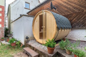 A wooden sauna structure with a glass door, surrounded by potted plants and a stone bench.