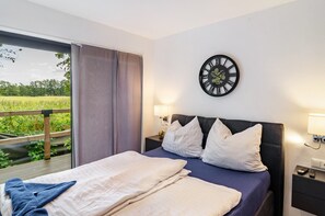 A bedroom with a bed, pillows, a wall clock, bedside lamps, and a window with a view of greenery.