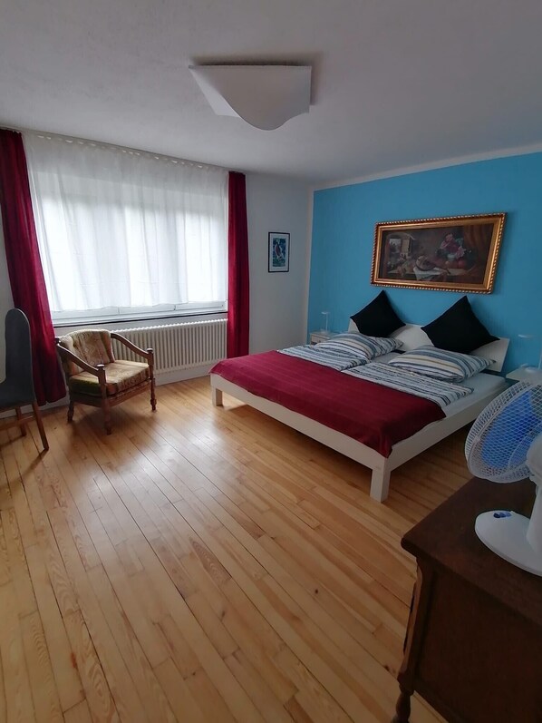 Ein Schlafzimmer mit Bett, Stuhl, Heizkörper, einem Fenster mit Vorhängen und einem Bild an der Wand.