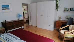 Une chambre à coucher comprenant un lit, une commode en bois, un bureau avec une chaise, une armoire et un ventilateur.