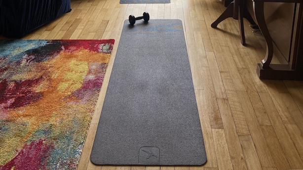 Une pièce avec un piano, une chaise bleue, un tapis coloré, un tapis de yoga et une vue sur l’extérieur, visible à travers une porte vitrée.