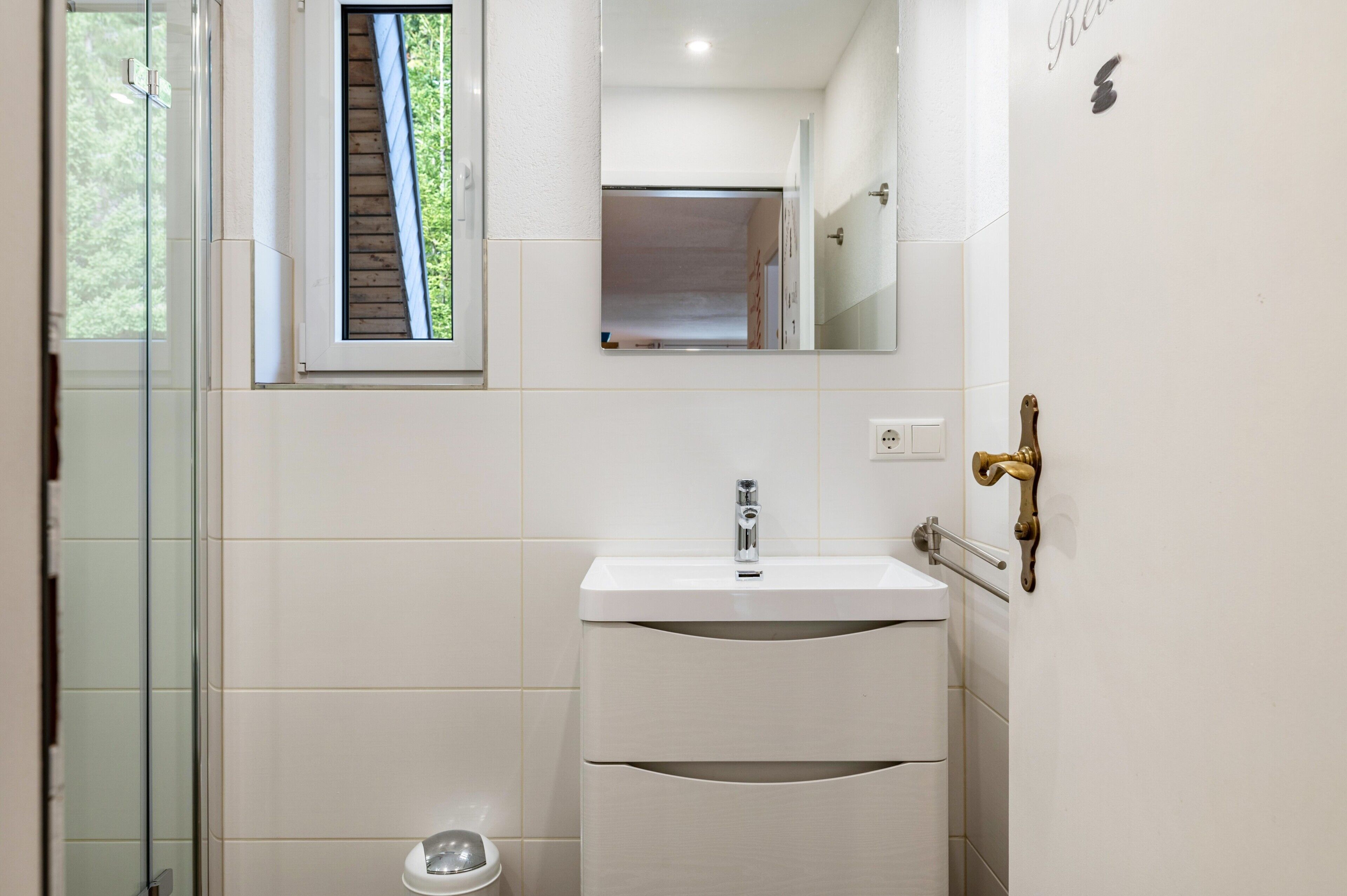 Un baño con cabina de ducha de vidrio, un lavamanos blanco con espejo y un picaporte de latón.