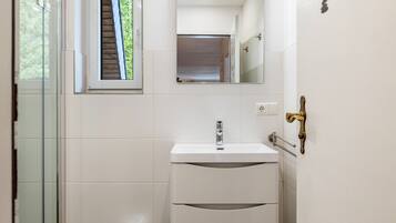 Un baño con cabina de ducha de vidrio, un lavamanos blanco con espejo y un picaporte de latón.