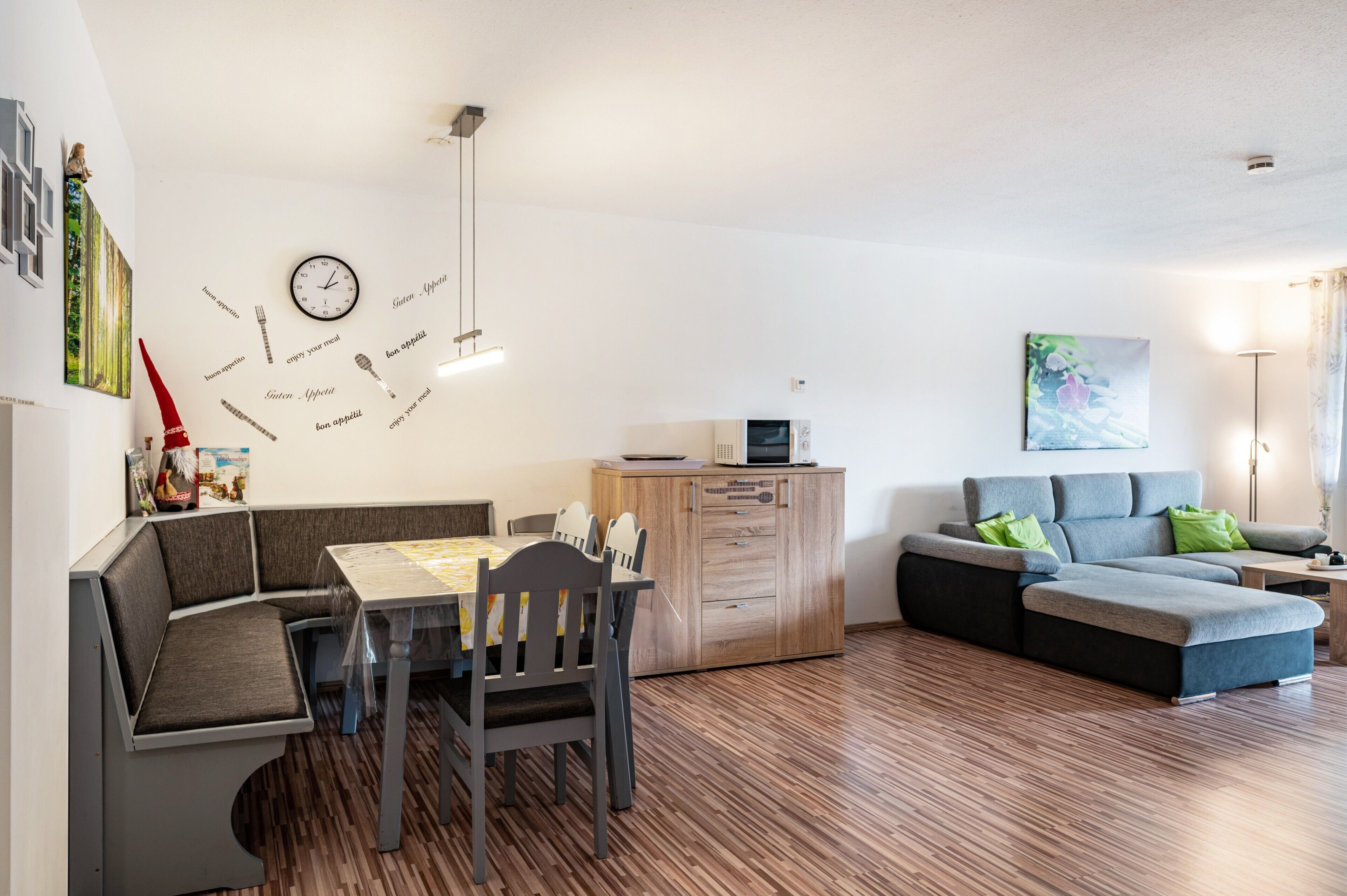 A modern living room with a dining area, a sofa, a wooden cabinet, and a wall clock.