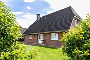 Une maison en briques avec un toit recouvert de tuiles sombres, une porte d’entrée et un petit jardin devant.