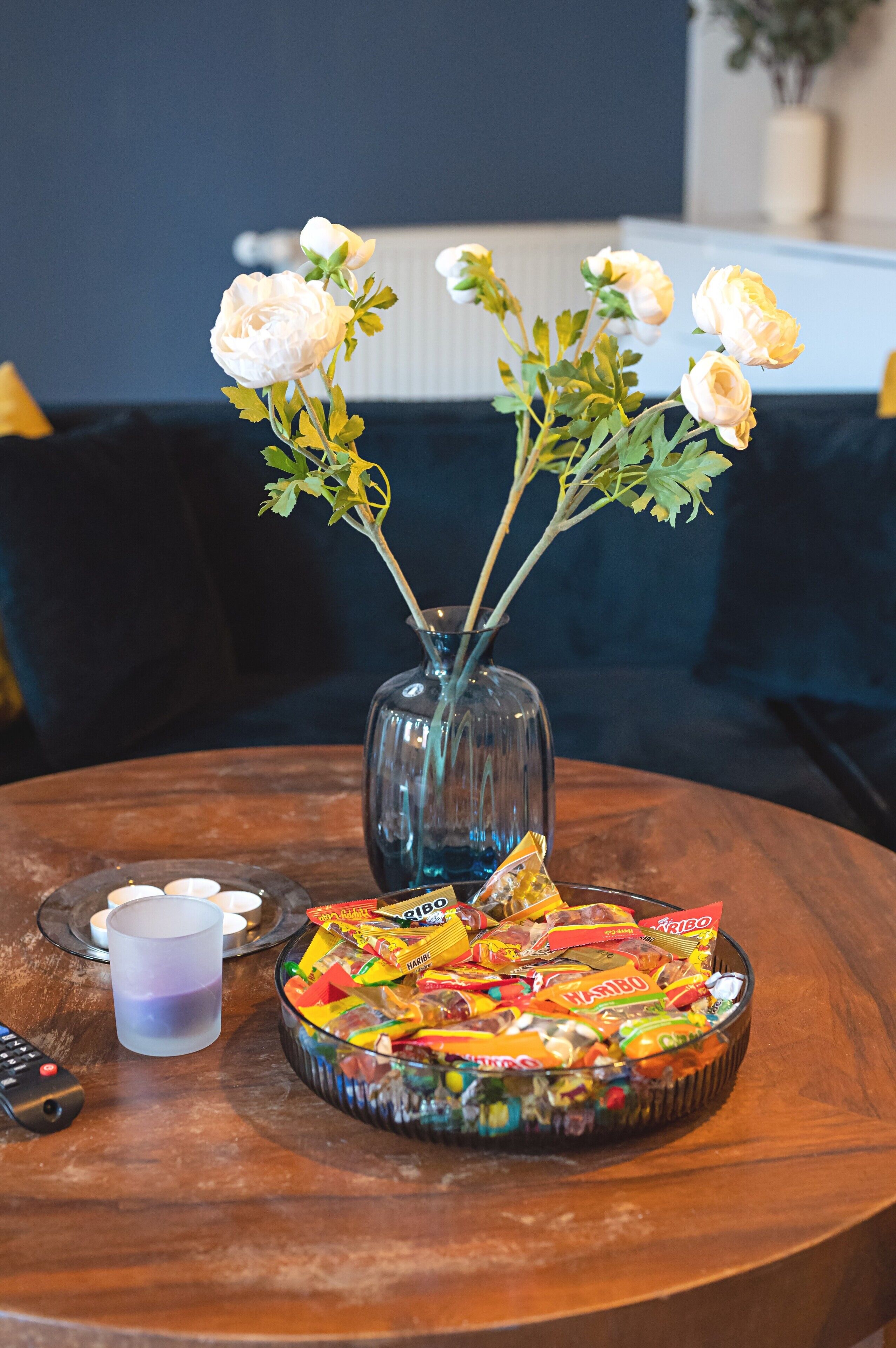 A round wooden table with a glass vase containing white flowers, a bowl of assorted candies, and a remote control.