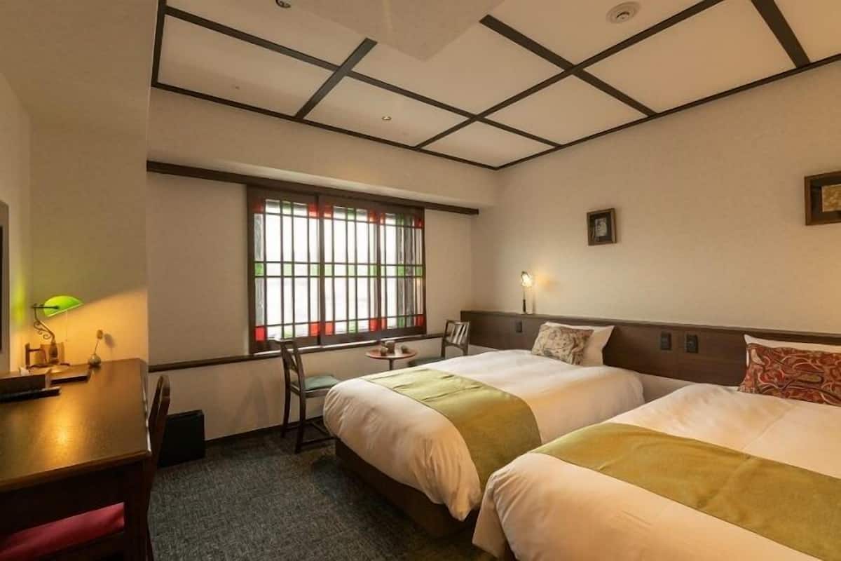 Habitación de hotel con dos camas, un escritorio, una silla, un ventanal con cortinas y una mesita.