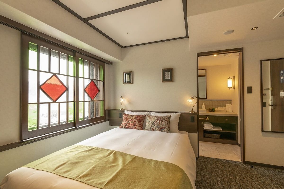 Habitación de hotel con cama, una ventana con vidrieras, espejo y baño visible a través de una puerta abierta.