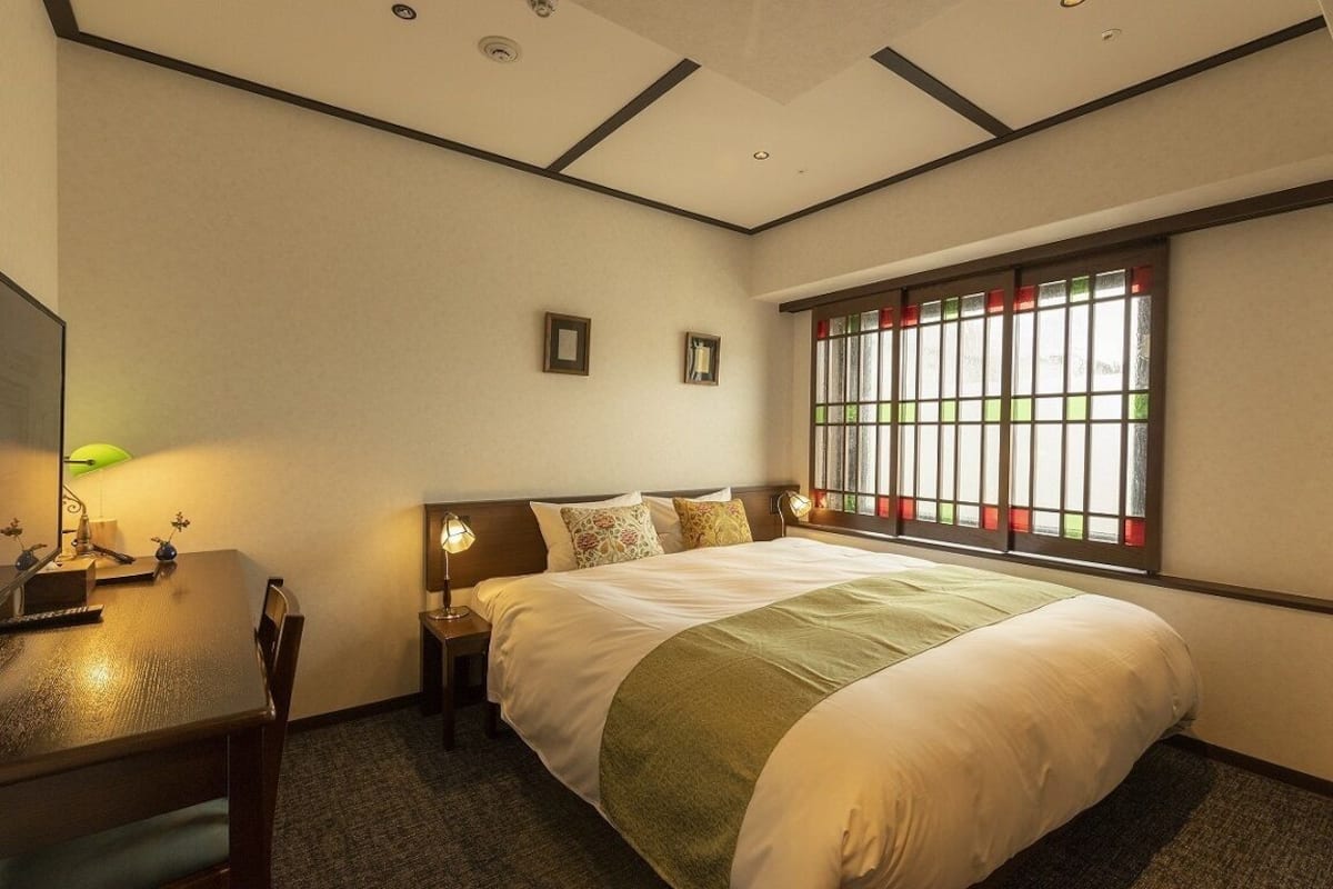 Habitación de hotel con una cama grande, un escritorio con lámpara, una ventana con paneles rojos y verdes, e iluminación empotrada en el techo.